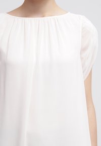 Top bianco con scollo plissettato e maniche corte trasparenti. Il tessuto è leggero e leggermente traslucido, con una texture liscia.