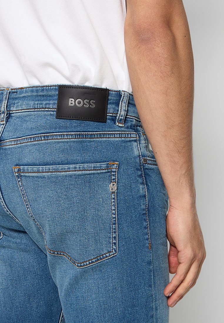 Jeans en denim bleu avec une patch en cuir noir arborant le logo "BOSS", design à cinq poches et accents de couture à l'arrière.