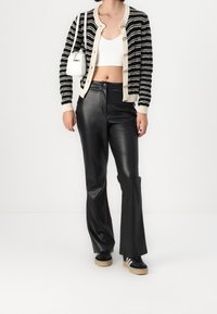 Kobieta w czarno-białym paskowanym kardiganie, białym topie crop, czarnych skórzanych spodniach, czarnych tenisówkach i niosąca białą torebkę na ramię.