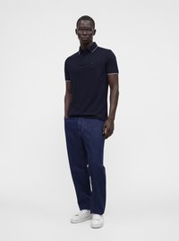 Homme portant un polo bleu marine foncé, un jean bleu, des baskets blanches et des lunettes, debout les mains dans les poches sur un fond clair uni.