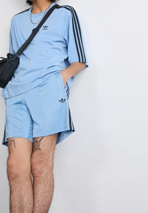 Osoba ubrana w jasnoniebieskie spodenki i koszulkę Adidas z czarnymi paskami, nosi czarną torbę przewieszoną przez ramię, stoi przy białej ścianie.