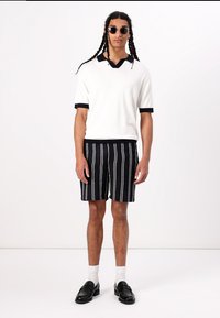 White short-sleeve polo with black accents, paired with black and white striped shorts. Model wears sunglasses, black loafers, and white socks.