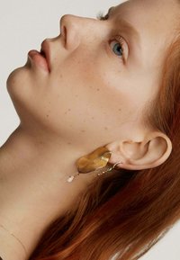 Pendientes en tono dorado con textura ondulada, que presentan un diseño de gota con acentos de cuentas transparentes y verdes. Ambientados en un fondo neutro.