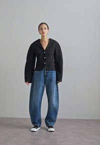 Black cropped blazer with a V-neck and large buttons, paired with wide-leg blue jeans and white sneakers, set against a neutral background.