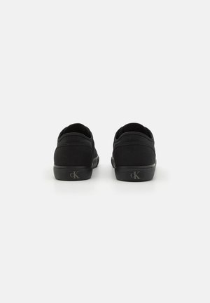 Par sorte lave sneakers med "cK" logo på hælen, vist bagfra på hvid baggrund.