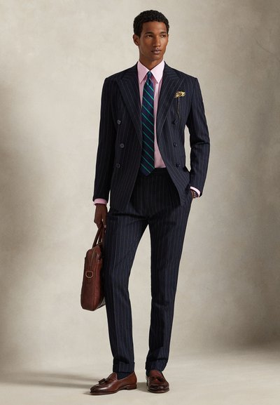 Férfi tengerészkék csíkos, kétsoros öltönyben, rózsaszín inggel, kék-zöld csíkos nyakkendővel, barna bőr aktatáskát tartva, bézs háttér előtt áll.