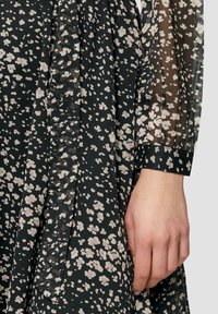 Schwarzes Blumenkleid aus einem weichen, durchsichtigen Stoff mit rosa- und weißen Blumenmustern. Die Ärmeln haben einen taillierten Bund, der dem Design Struktur verleiht.