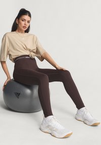 Brown high-waisted leggings with white side accents, paired with a beige knotted t-shirt and white athletic shoes, sitting on a grey exercise ball.