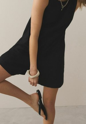 Femme en robe noire sans manches portant un gros bracelet blanc, tenant un pied avec une sandale noire partiellement enlevée.