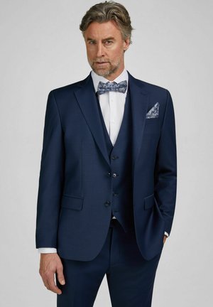 Mann trägt einen marineblauen Dreiteiler mit weißem Hemd und gemusteter blauer Fliege, posiert mit einer Hand in der Tasche vor grauem Hintergrund.