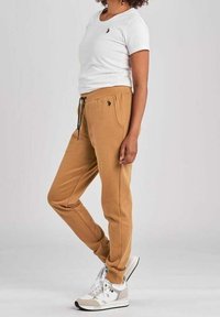 Camicetta bianca a maniche corte con un piccolo logo nero, abbinata a pantaloni joggers beige con coulisse e tasche laterali, indossati con sneakers bianche.
