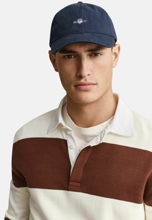 Giovane uomo che indossa un cappellino GANT blu navy e una maglietta polo bianca con una larga striscia orizzontale marrone, guarda dritto davanti a sé.