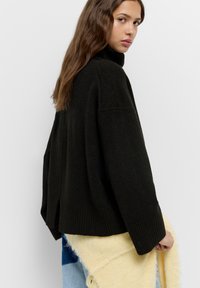 Črn, prevelik pulover s turtleneck ovratnikom in ribastim robom ter stranskimi razpoki. Pletenina izgleda mehka in udobna, prosto pada.