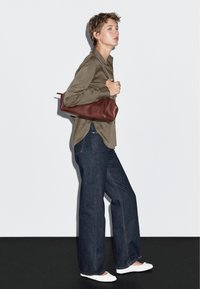 Camicia verde oliva a maniche lunghe, jeans larghi blu scuro, scarpe bianche slip-on e una borsa a tracolla in pelle marrone su uno sfondo neutro.