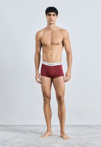 Czerwone slipy z wzorem w kratkę, z białym pasem w talii z logo "Calvin Klein". Gładki materiał i dopasowany krój. Model stoi boso.
