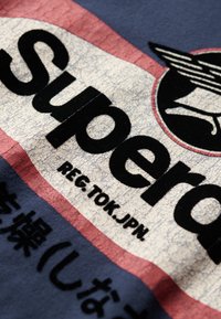 Camiseta gráfica con un fondo de color crema agrietado y texto negro en negrita "Super" con acentos rojos; fabricada principalmente en tela de color azul marino.