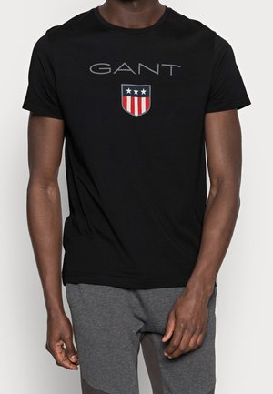 T-shirt en coton noir avec col rond, manches courtes, et logo gris "GANT" avec un design de blason comportant des rayures rouges et blanches et des étoiles bleues.