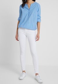 Blouse en coton bleu clair avec des rayures verticales vertes, manches courtes retroussées, associée à un jean skinny blanc et des baskets blanches.