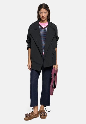 Młoda kobieta ubrana w czarną oversize kurtkę, pasiasty top, ciemne dżinsy, brązowe mokasyny, trzymająca w jednej ręce złożoną fioletową torbę.