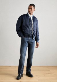 Marine bomberjakke med glidelås foran, ribbede mansjetter og hem, brukt over en hvit skjorte og kombinert med mellomblå jeans og svarte støvler.