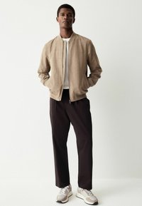 Tan suedebomberjakke med lynlås foran, parret med sorte bukser og tan-grå sneakers. Modellen står med hænderne i lommerne mod en lys baggrund.