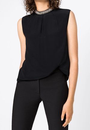 Vrouw draagt een mouwloze zwarte top met een met kralen versierde kraag en zwarte broek, staand met één hand in de zak tegen een effen achtergrond.