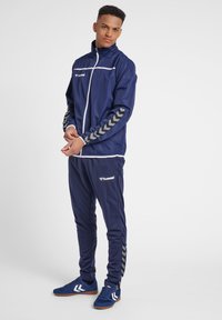 Marinblå träningsoverall i lättviktigt material, med vita accenter, hel dragkedja och fiskbensmönster på ärmarna och byxorna.