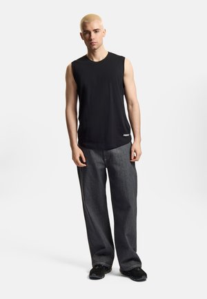 Młody mężczyzna o krótkich blond włosach, ubrany w czarny bezrękawnik, luźne ciemnoszare dżinsy i czarne trampki, stojący na białym tle.