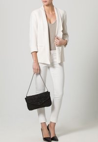 Bílý blazer přes béžový top, doplněný bílými kalhotami. Černá kabelka s řetízkem a špičaté černé lodičky dotvářejí celkový vzhled.