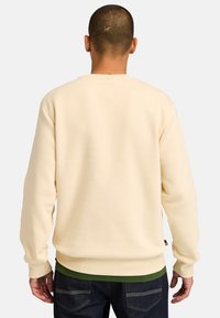 Sweatshirt crème avec des poignets et un ourlet côtelés, doté d'un col rond. Porté par-dessus une chemise verte, le tissu semble doux et lisse.