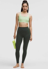 Világos zöld sportmelltartó és sötétzöld, magas derekú leggings, mindkettő rugalmas anyagból készült. Egy fényeszöld vízpalackot tartanak az egyik kezükben.