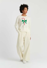 Maglione crema con un fiocco verde e un design a bastoncino di zucchero a strisce rosse, abbinato a pantaloni larghi di colore chiaro e sneaker bianche.