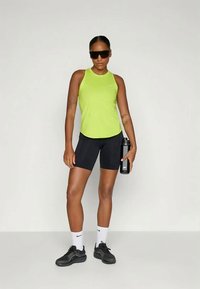 Haut de sport sans manches vert vif, shorts ajustés noirs, chaussures de sport noires, chaussettes blanches et lunettes de soleil noires ; tenant une bouteille d'eau.