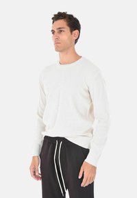 Maglione bianco a coste con maniche lunghe e scollatura rotonda, indossato sopra pantaloni neri con coulisse. Il tessuto appare morbido con una finitura strutturata.