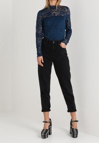 Top in pizzo blu navy a maniche lunghe con motivo floreale, abbinato a pantaloni neri a vita alta e scarpe con tacco a piattaforma a scacchi bianchi e neri.