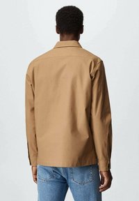 Chemise marron à manches longues en tissu texturé avec un col classique et des boutons en détails. Portée avec un jean en denim bleu clair.