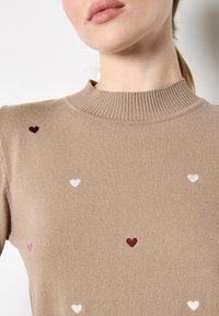 Beige trui met korte mouwen, een geribbelde halslijn en verspreid geborduurde hartjes in bordeauxrood, roze en wit. Zachte, gestructureerde brei stof.