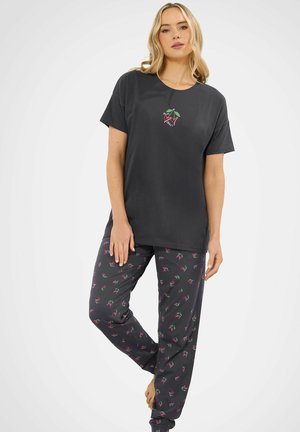 Donna che indossa una maglietta nera a maniche corte con una piccola grafica di ciliegie e pantaloni neri abbinati con motivo di ciliegie rosa e verdi, in piedi scalza.