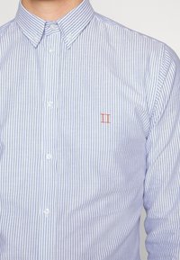Camicia a maniche lunghe in cotone blu chiaro e bianco a righe. Presenta un colletto classico e un piccolo logo rosso sul petto.