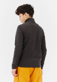 Donkergrijze fleece pullover met hoge kraag en lange mouwen, gecombineerd met felgele broek, gezien vanuit de achterkant.