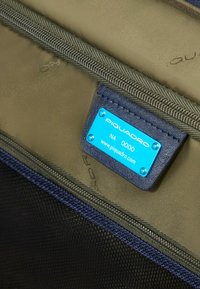 Strukturierte olivgrüne Innenausstattung mit einem marineblauen Reißverschluss. Blauer Metallanhänger mit Prägung "PIQUADRO NA 0000" und Website.