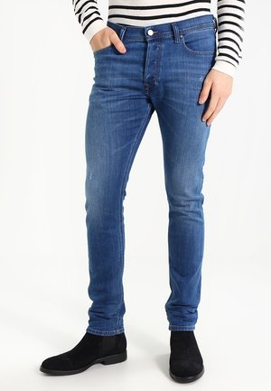 Mand står iført slim fit blå jeans, sorte ruskindssko og en hvid langærmet skjorte med sorte horisontale striber.