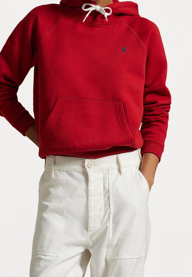 Felpa rossa con tasca frontale e cordoncino bianco, caratterizzata da un piccolo logo blu. Abbinata a pantaloni bianchi a vita alta con texture.