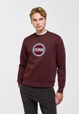 Młody mężczyzna w bordowej bluzie z okrągłym logo "Colmar", stoi z jedną ręką w kieszeni, lekko patrząc na bok.