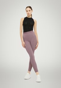 Zwarte mouwloze bodysuit van textuurstof, gecombineerd met lichtpaarse leggings met een patroon op de tailleband en zijdetails, aangevuld met witte sneakers.