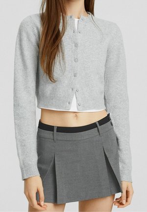 Femme portant un cardigan court gris clair sur un haut blanc et une mini-jupe plissée grise taille haute avec un détail de ceinture noire.