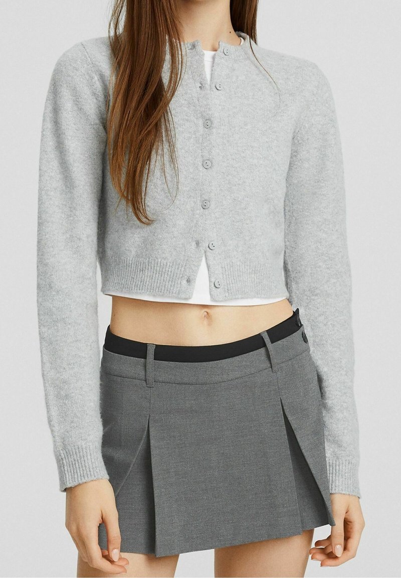 Femme portant un cardigan court gris clair sur un haut blanc et une mini-jupe plissée grise taille haute avec un détail de ceinture noire.
