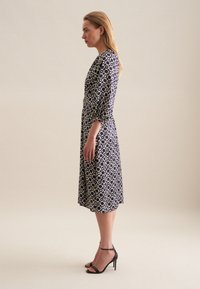Granatowa sukienka z kremowym geometrycznym wzorem, z rękawami długości trzech czwartych i zwiewną sylwetką. Czarne sandały na obcasie dopełniają stylizację.