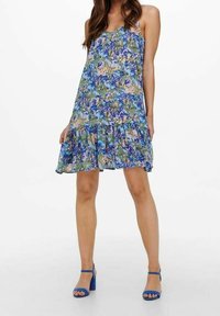 Kleid mit Blumendruck in Blau, Grün und Pink, mit verstellbaren Trägern und einem geschichteten Rüschensaum. Kombiniert mit blauen Blockabsatzsandalen.