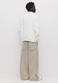 Overmaat witte sweatshirt met een hoge kraag, gecombineerd met losse beige broek met twee achterzakken, gepresenteerd tegen een effen achtergrond.